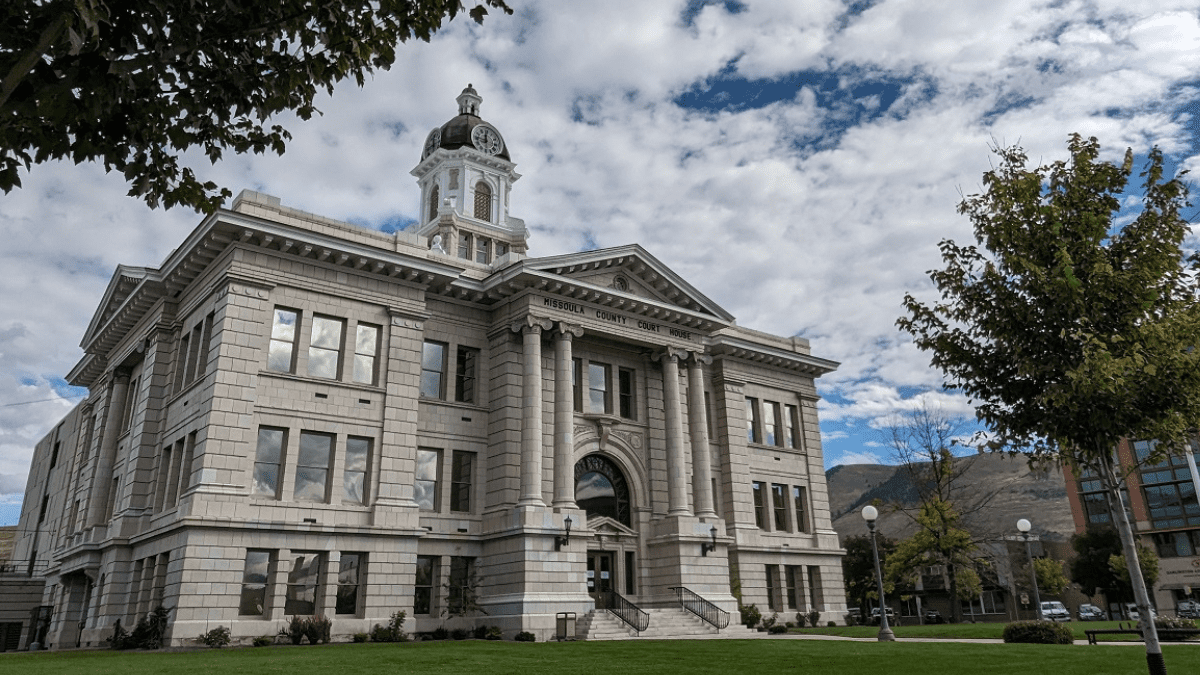 The Missoula County Courthouse surrounded by trees and a green lawn under a partly cloudy sky.