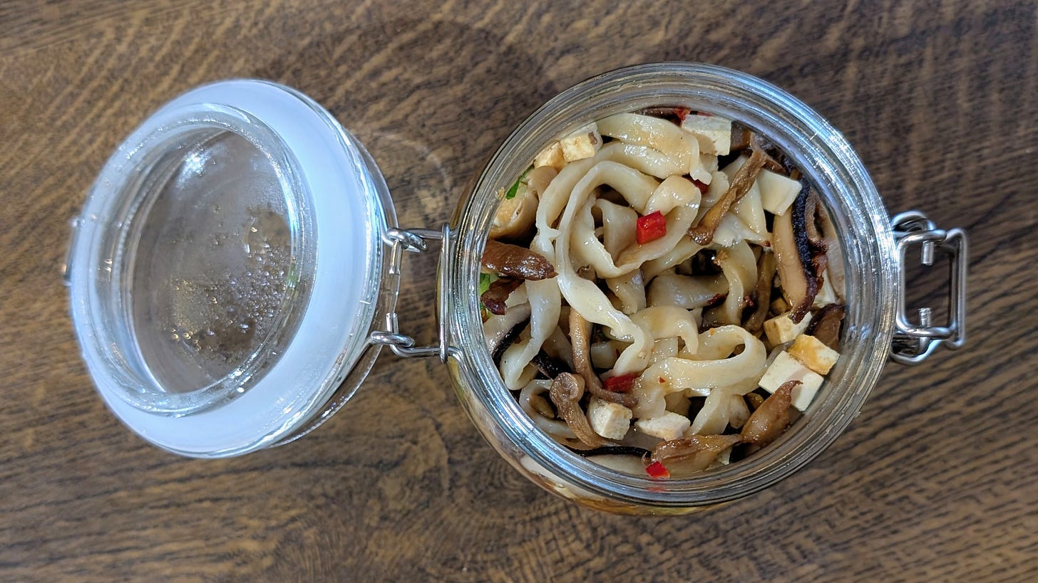 An open glass jar with a hinged lid sits on a wooden surface, filled with flat noodles, diced tofu, sliced mushrooms, red chilli pieces, and green herbs. Condensation clings to the inside of the jar, making the layered ingredients look warm and freshly prepared.