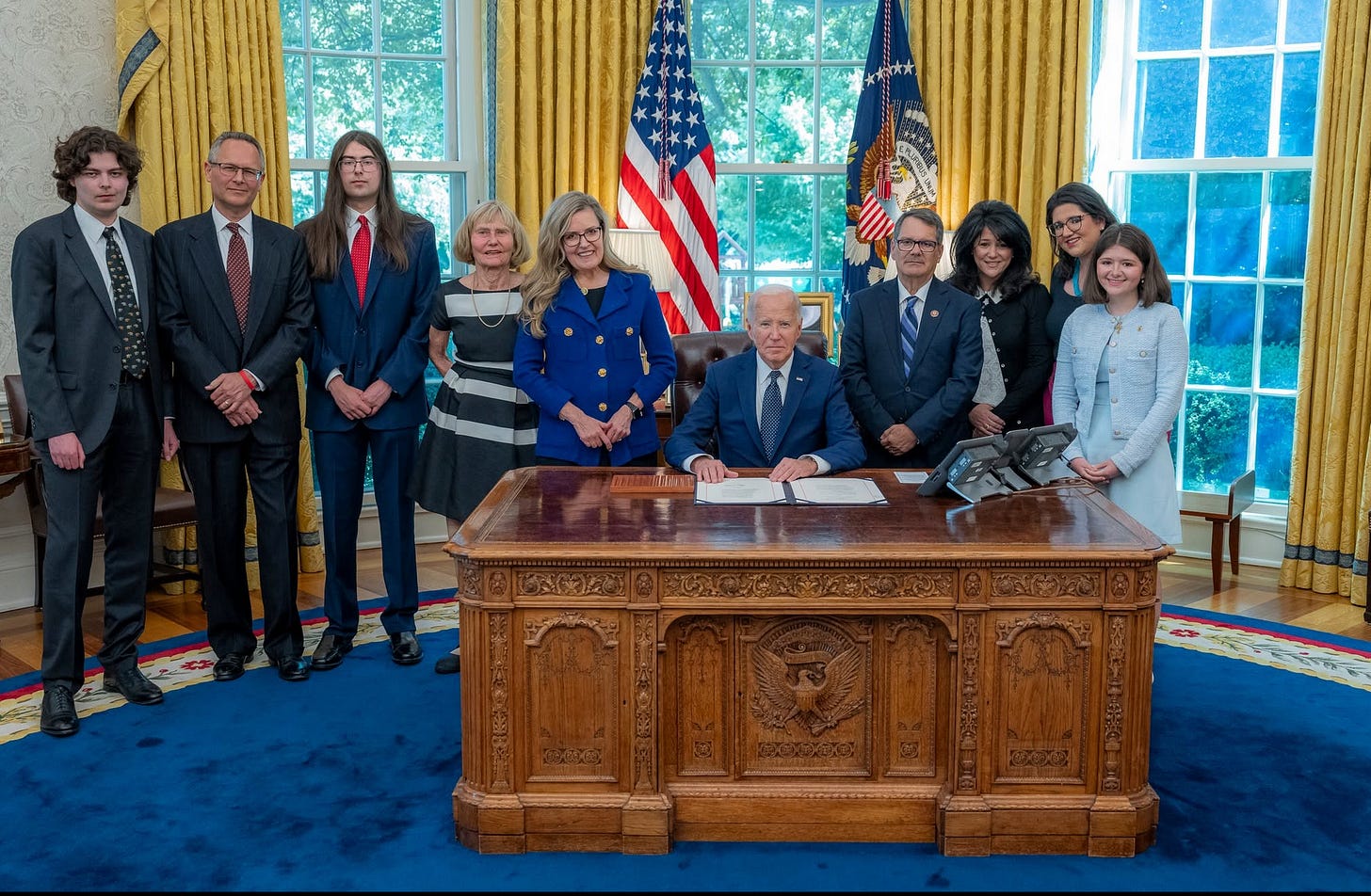 President Biden seated at the Resolute Desk signing a bill, surrounded by a group of standing guests.
