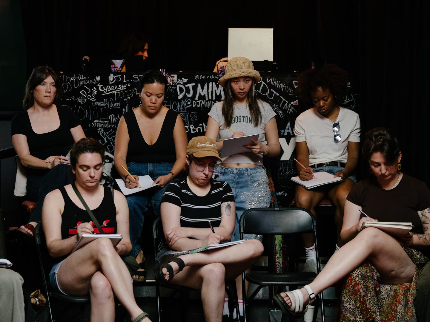 A group of people sit closely together at a bar-hosted figure drawing event, sketching a nude model off-camera while holding clipboards and notebooks. A group of people sit closely together at a bar-hosted figure drawing event, sketching a nude model off-camera while holding clipboards and notebooks.