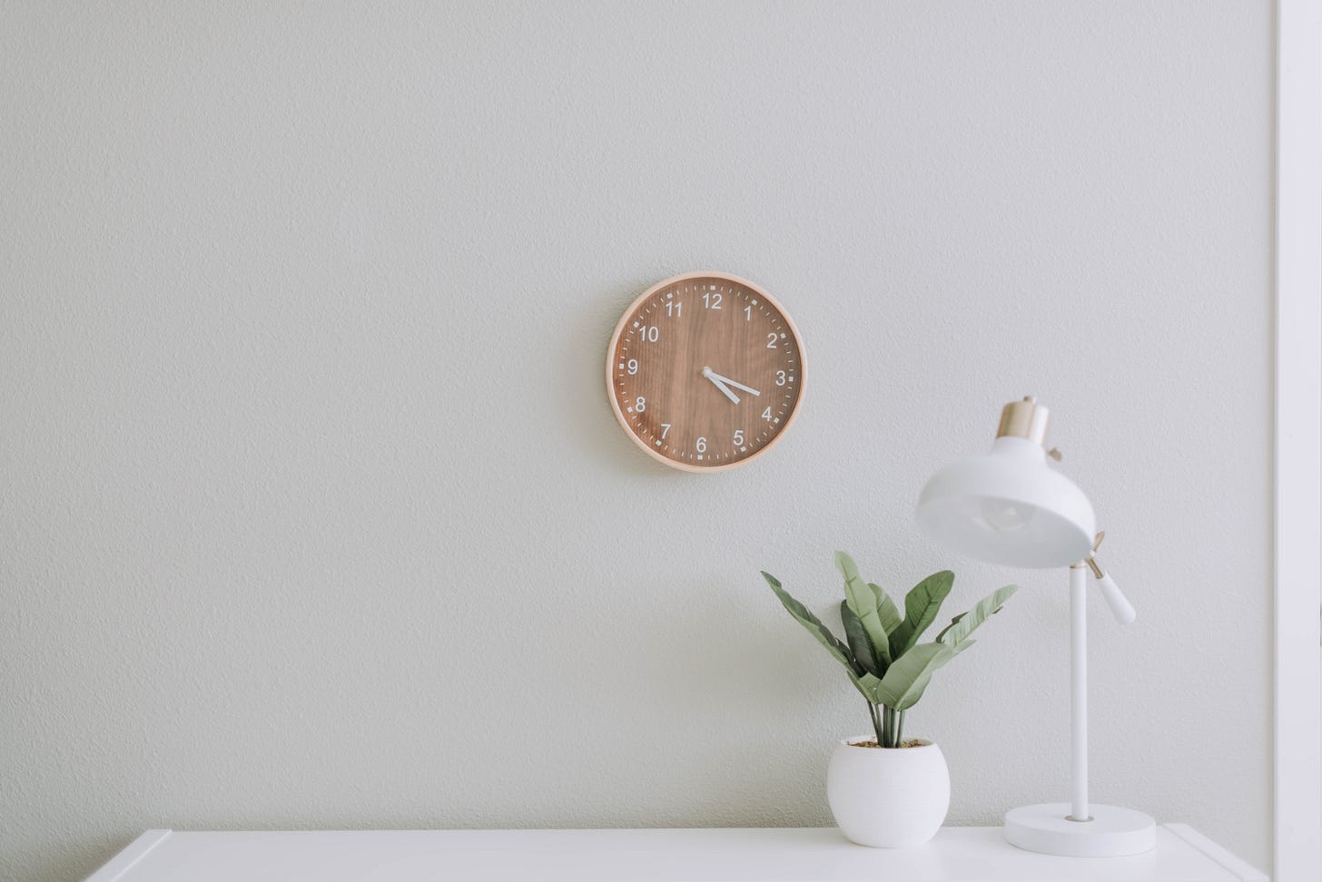 a white desk with a plant and lamp on top and a brown clock on the wall a white desk with a plant and lamp on top and a brown clock on the wall