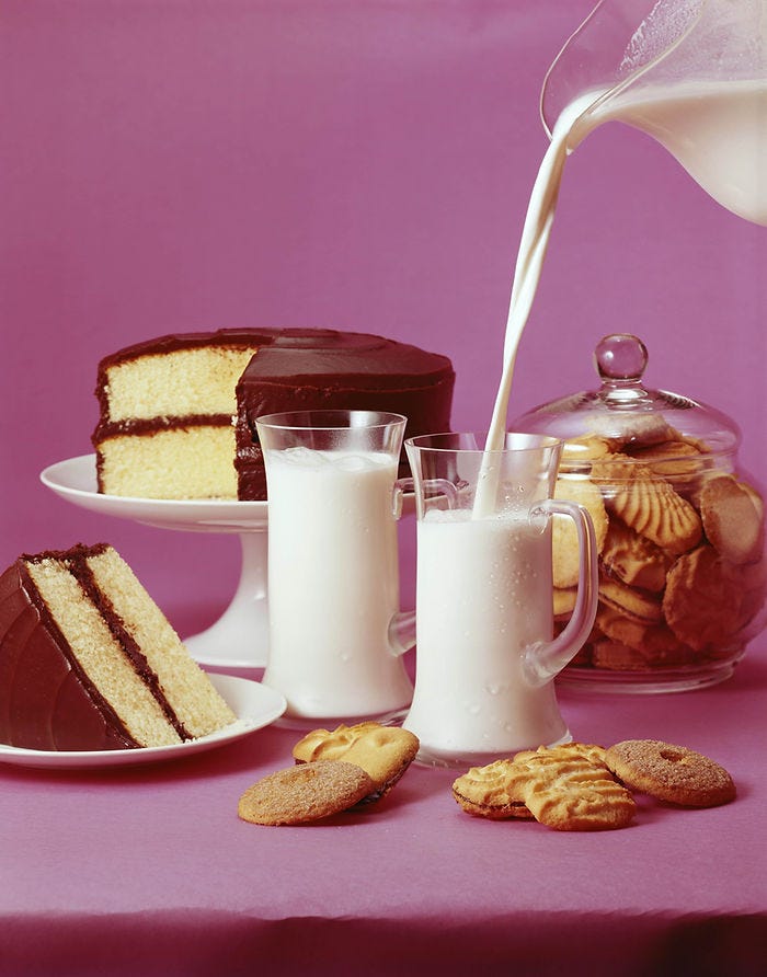 Chocolate cake and milk served on a purple table with cookies. Milk is being poured into glasses, creating a calm, inviting scene.