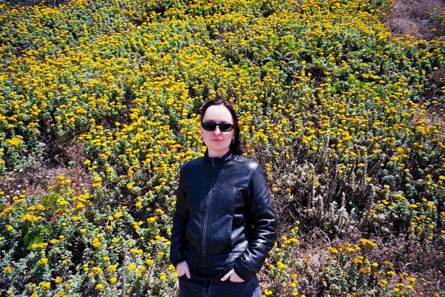 Evyn Silver, a white femme with long dark hair, stands with her hands in her pockets in front of a hill covered in bright yellow flowers. She wears black sunglasses, a leather jacket, and dark jeans.