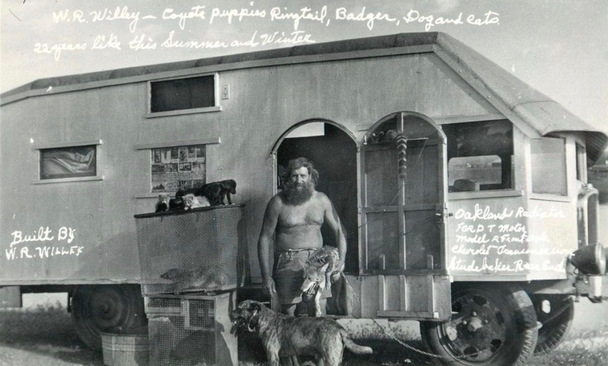 Black-and-white photograph of Willie Willey standing bare-chested in shorts outside a homemade trailer mounted on a vehicle chassis, with several animals including dogs and small mammals gathered around him. Handwritten notes on the trailer identify Willey as the builder and list the vehicle’s mixed automotive parts, capturing his traveling home during his 1951 stop near Walla Walla.