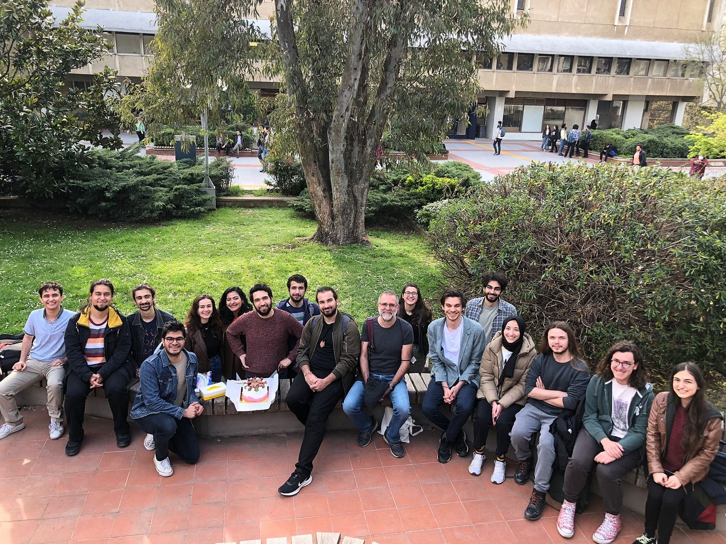 15-20 kadar öğrenci basamak gibi bir yere oturmuşlar, ortalarında hocaları. Herkesin yüzünde çok güzel bir gülümseme. 