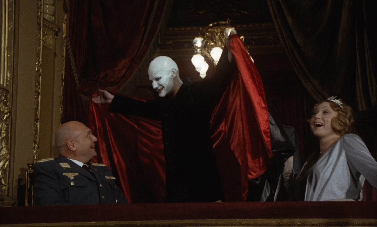 In a box at the theatre where he's performing, actor Hendrik Höfgen (Klaus Maria Brandauer), made up as Mephistopheles (complete with bald head covered in white make-up) unfurls  his resplendent cloak in front of the General (Rolf Hoppe) and Lotte Lindenthal (Christine Harbort).