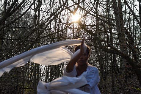 Me, a Black woman wearing a floaty white top is dancing with a white voile which billows around her. The weak Spring sunlight flares through the trees in the woodland behind her.