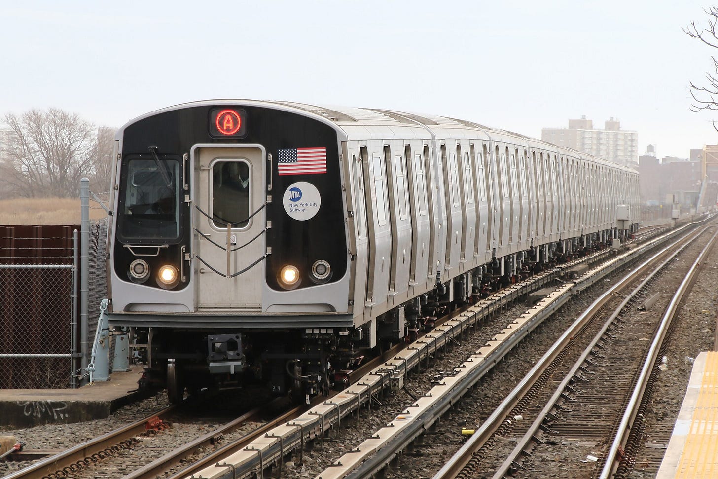 An A train, composed of R179 cars, is seen entering the Broad Channel station. The front of the train contains two red tail lights, a window on the left side, the American flag on the right side, and the MTA logo below the flag.