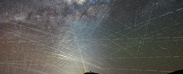 The background of this image shows a starry sky, including the bright lane of the Milky Way. Hundreds of bright streaks cross the sky in different directions, as if scratching the background natural sky behind it. The bottom of the image is occupied by the dark silhouette of a mountainous desert landscape. Atop the mountain at the centre, is a small, distant metallic dome and yellow-orange lasers shine behind it. The background of this image shows a starry sky, including the bright lane of the Milky Way. Hundreds of bright streaks cross the sky in different directions, as if scratching the background natural sky behind it. The bottom of the image is occupied by the dark silhouette of a mountainous desert landscape. Atop the mountain at the centre, is a small, distant metallic dome and yellow-orange lasers shine behind it.