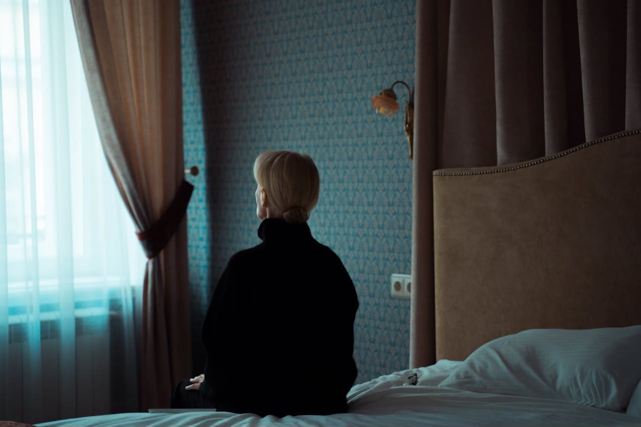 Back view of a mature woman wearing a black turtleneck while sitting on the edge of her bed and looking out the window. Back view of a mature woman wearing a black turtleneck while sitting on the edge of her bed and looking out the window.