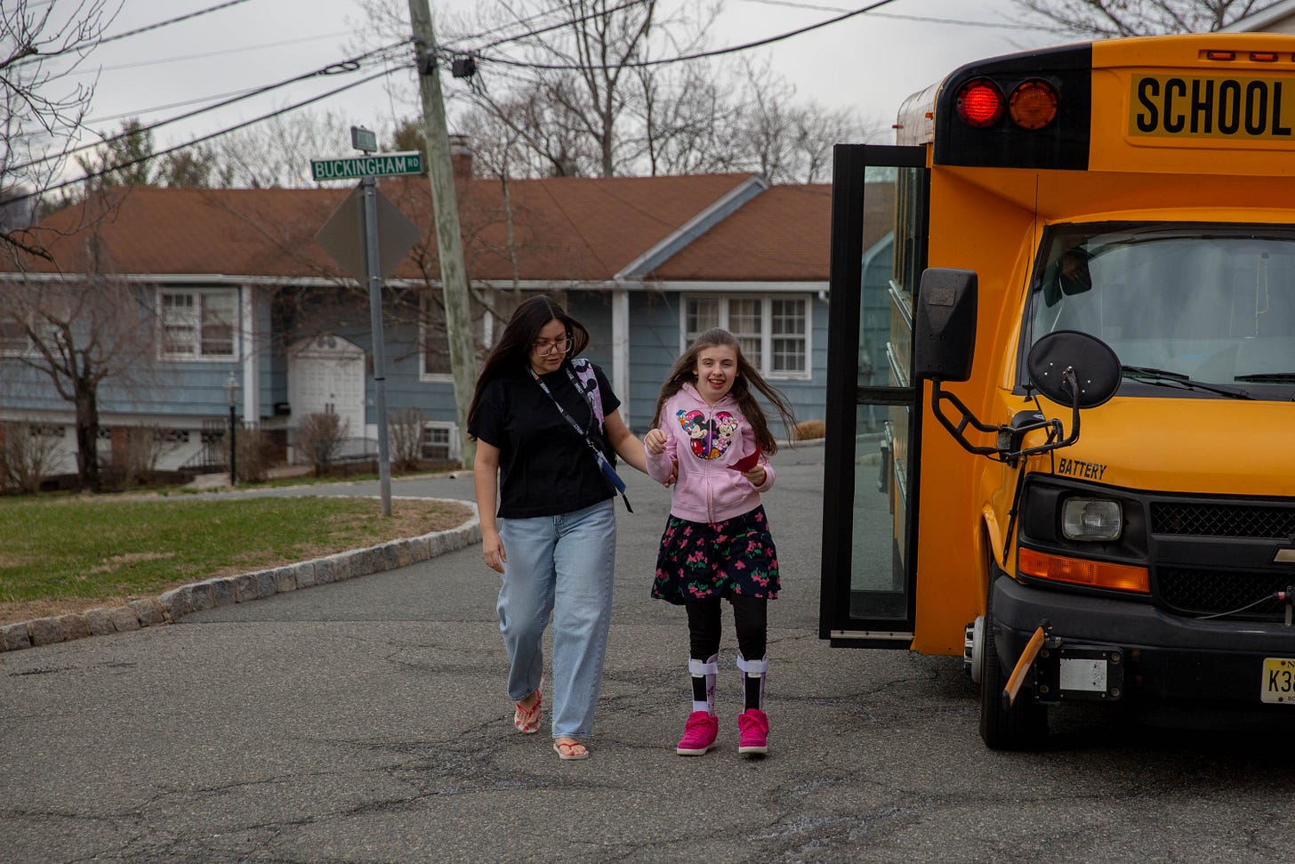 A woman with long brown hair wearing glasses walks away from a school bus with a girl wearing a pink sweatshirt and flowered dress A woman with long brown hair wearing glasses walks away from a school bus with a girl wearing a pink sweatshirt and flowered dress