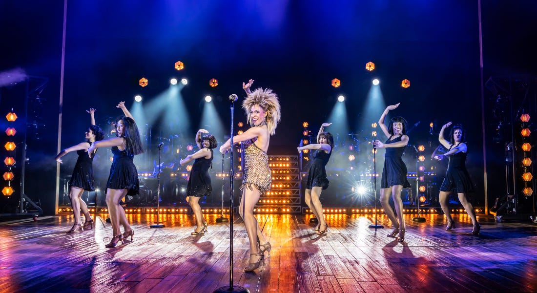 A production image from Tina The Tina Turner Musical with a woman standing on a stage in a gold dress and backing dancers in black dressers behind her