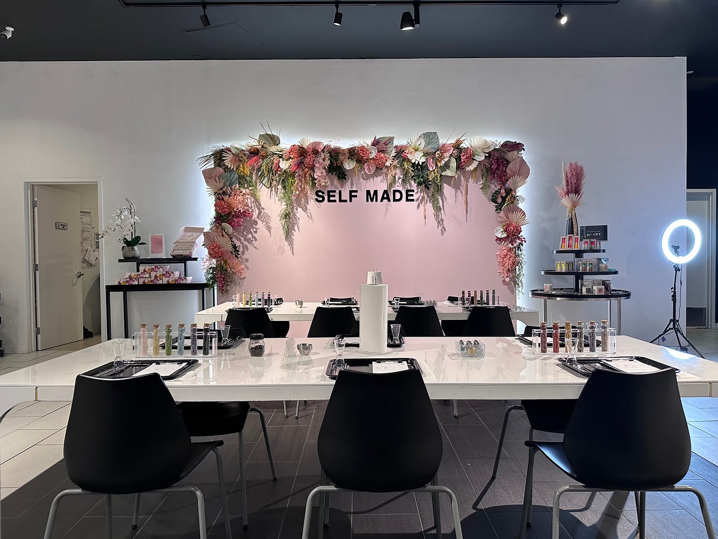 A stylish candle-making workstation at Blank Mason featuring a floral-decorated backdrop wall with 'SELF MADE' text in bold black letters. The wall has a pink background adorned with dried flowers in pink, white, and burgundy tones. In front is a clean white workstation with black chairs where customers can create their candles. Ring lights and candle-making supplies are visible, all designed for an Instagram-worthy experience.