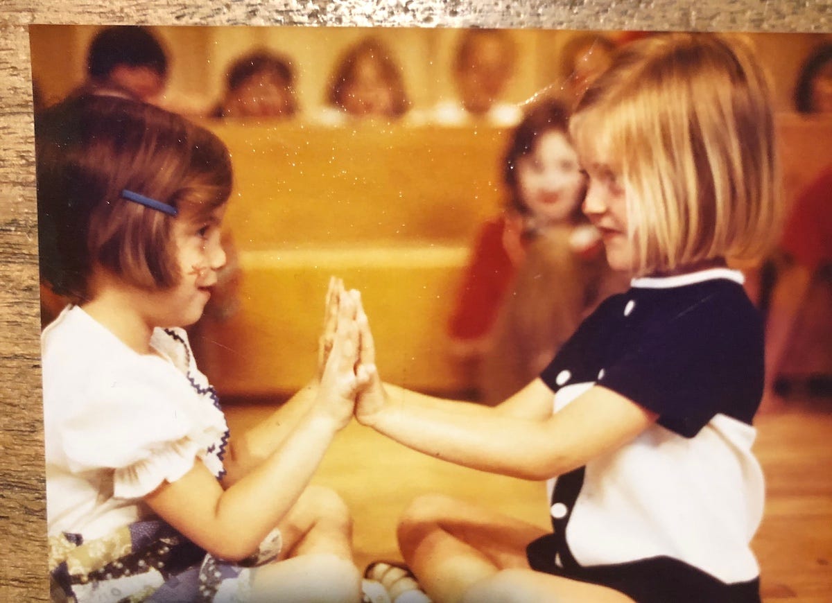 Two little girls sit facing each other, touching hands, playing patty-cake.