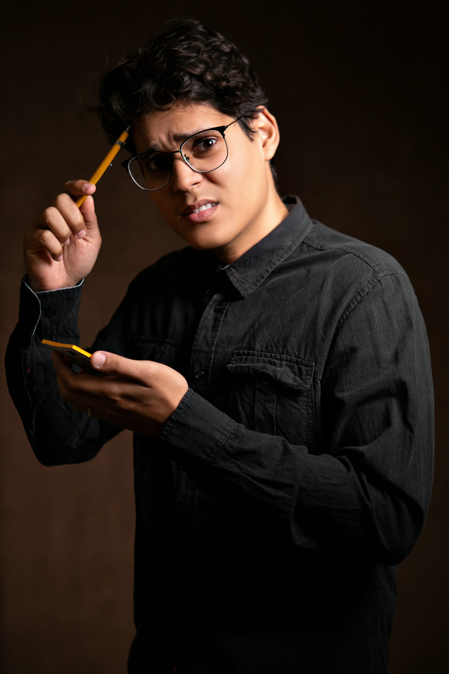Young creative professional with glasses holding pencil and smartphone, thoughtful expression in dark artistic lighting