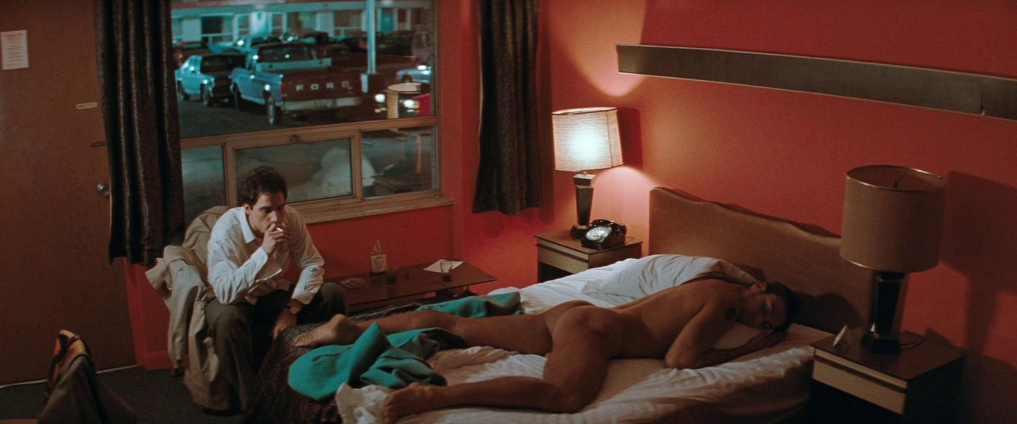 In a reddish motel room with its blinds open, Elias Koteas sits smoking a cigarette, fully clothed but looking disheveled. A nude man sleeps on the bed just in front of him, which is in a state of disarray. In a reddish motel room with its blinds open, Elias Koteas sits smoking a cigarette, fully clothed but looking disheveled. A nude man sleeps on the bed just in front of him, which is in a state of disarray.