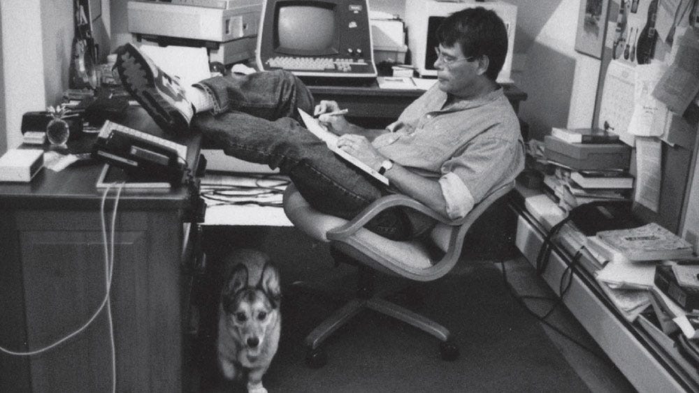 Stephen King writing at his desk with his dog underneath.