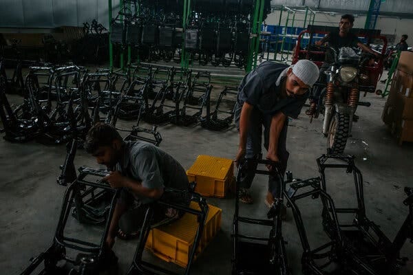 Two people work on black metal motorcycle frames lined up in rows on a concrete floor.