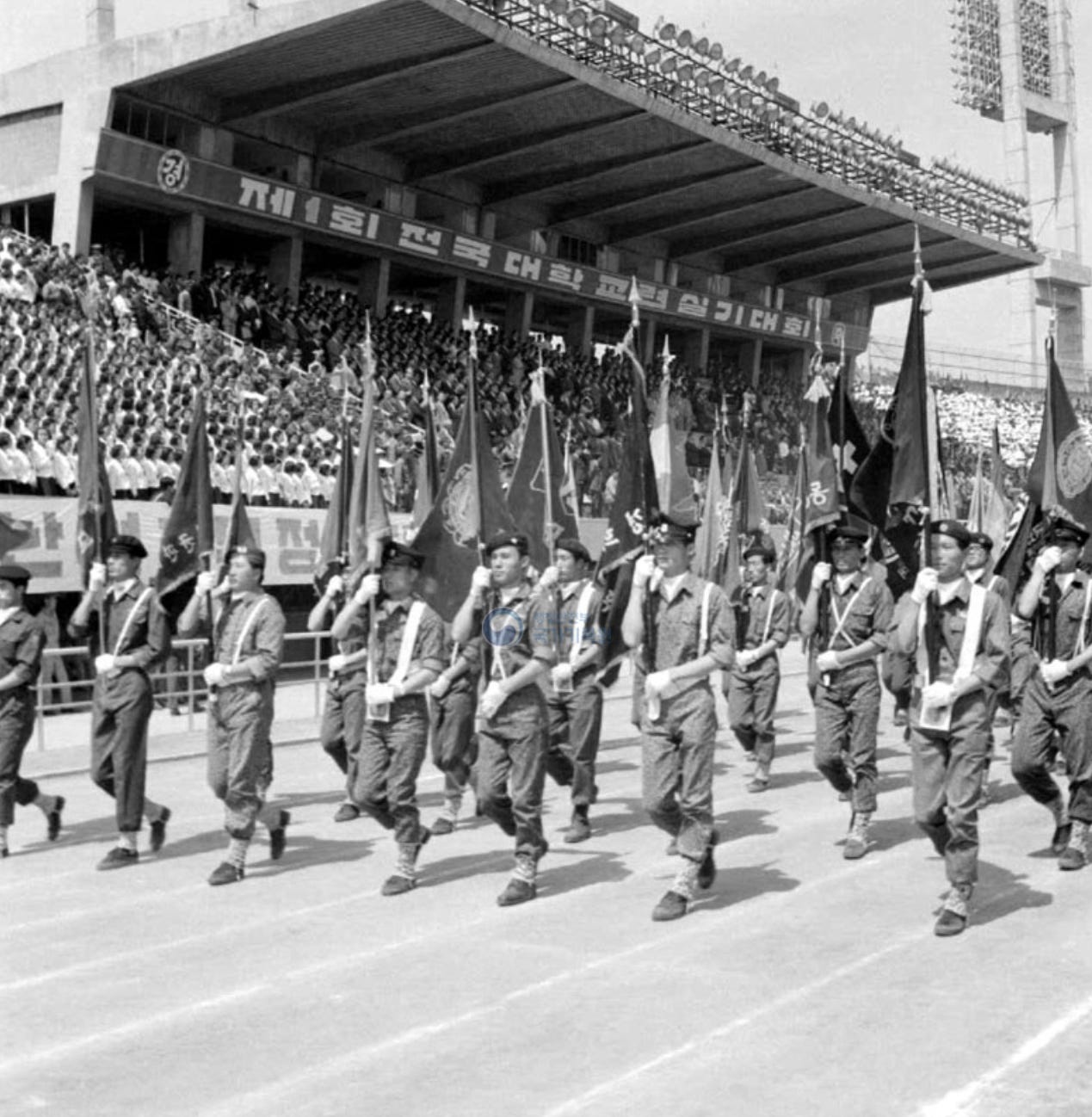 Black-and-white photo, 1969: university students in fatigues and caps march in formation on a stadium track, each row carrying large unit flags; the stands are full and a banner overhead reads “1st National University Military Training Skills Competition” in Korean.