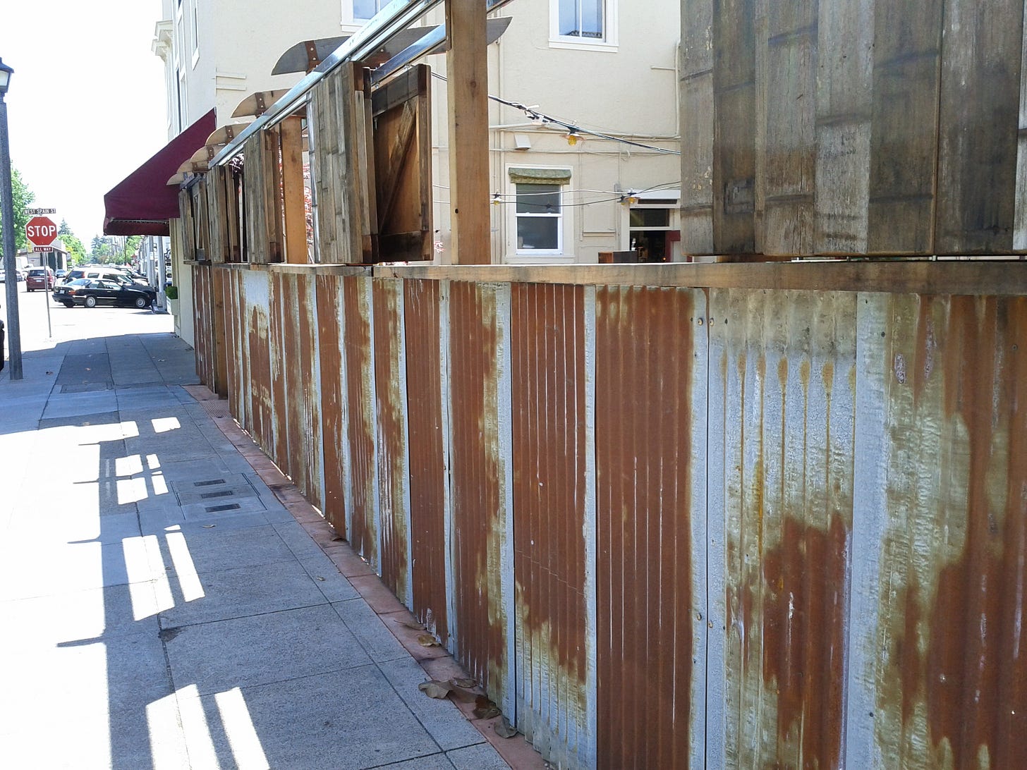 A sidewalk in a small town on a sunny day, lined by a rusty old corregated fence