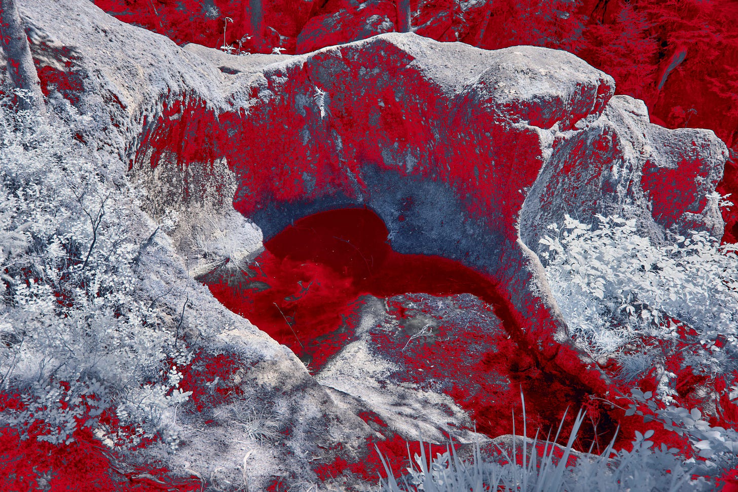 An infrared photograph of a rocky landscape, colours inverted: shadows rendered deep arterial red, stone bleached to pale grey-white. At the centre, a natural hollow in the rock forms a dark opening, pooling into deeper red shadow. 