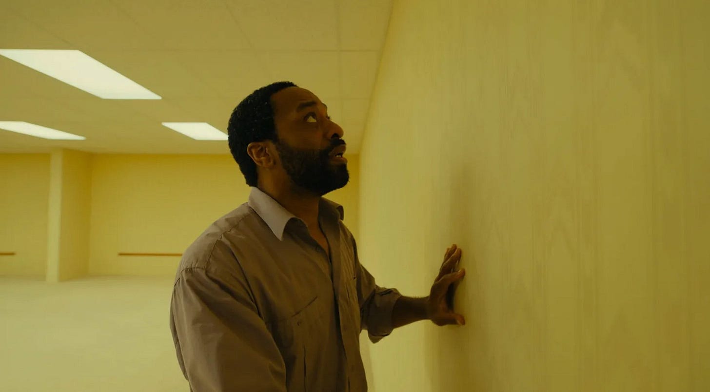 Still shot from the movie "Backrooms" with yellow walls. Clark presses hand against wall