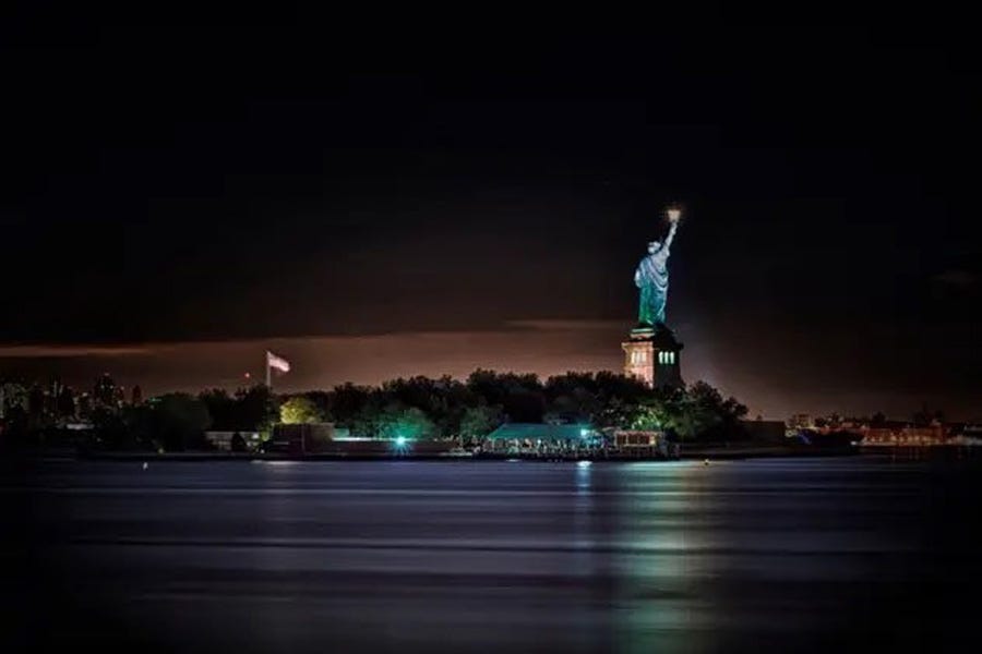 Nighttime photo of Liberty Island, with the Statue of Liberty, seen from behind, brightly lit, the colors of the green statue and gold torch reflecting on the water. A huge US flag is also brightly lit in the darkness.