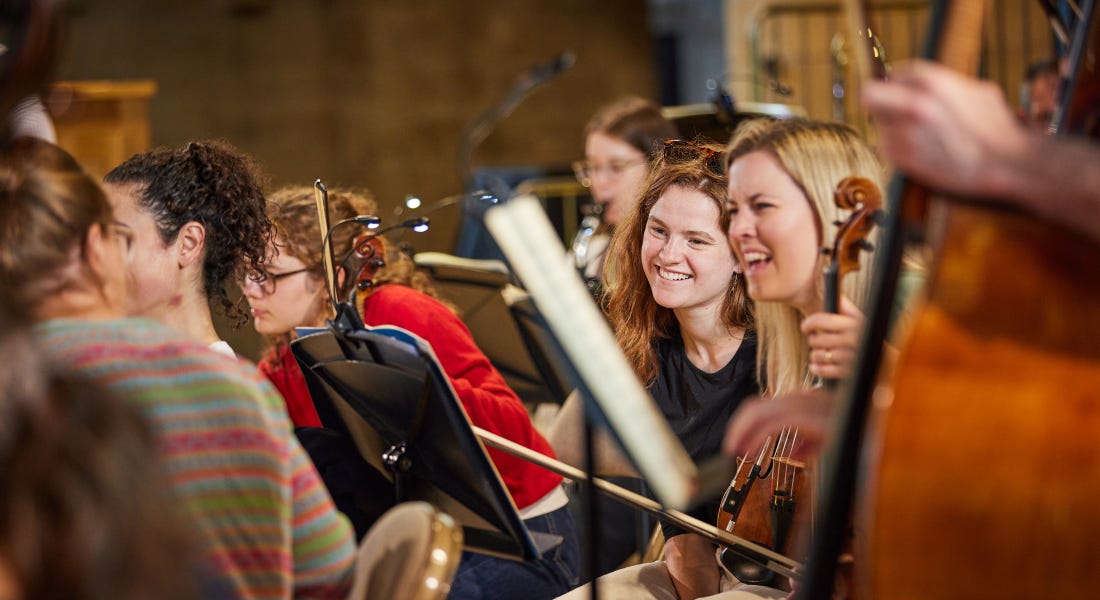 A close-up of an orchestra focusing on two women with violins smiling A close-up of an orchestra focusing on two women with violins smiling
