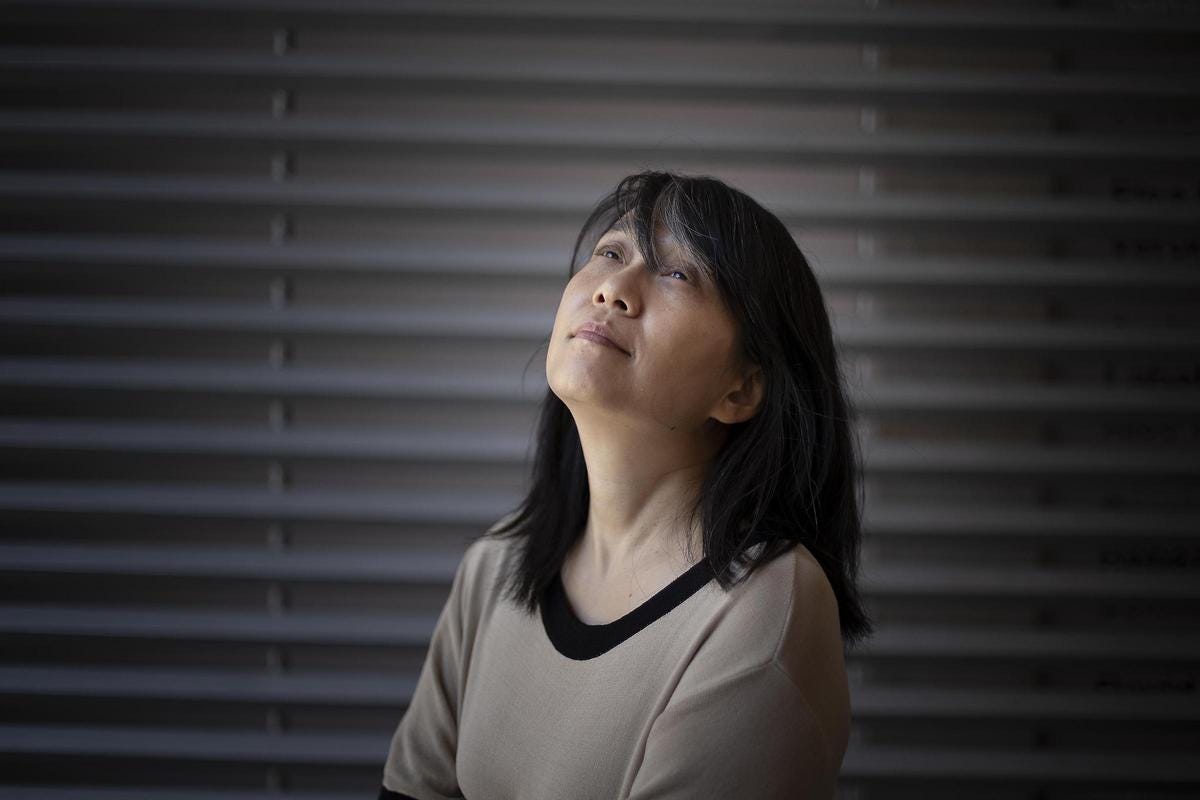 La imagen muestra a una mujer asiática con cabello oscuro y largo. Tiene una expresión calmada y serena, mirando directamente a la cámara. Viste una camiseta azul oscura y parece estar en un entorno con fondo gris o blanco que enfoca la atención en su rostro. Es una imagen retrato que captura la presencia tranquila y reflexiva de la mujer.