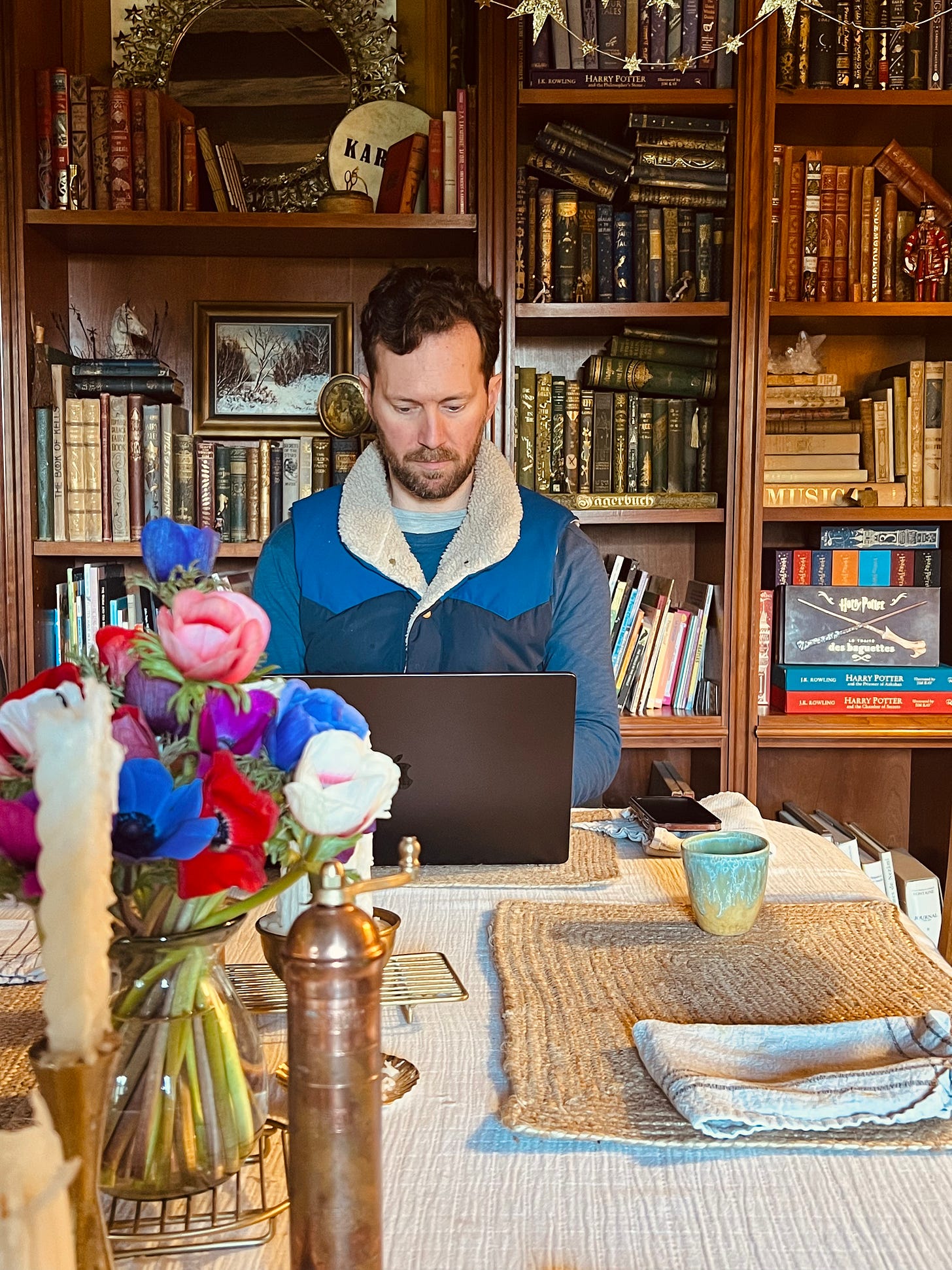 Sean working on a laptop at a kitchen table, with a vase of anemones in the foreground and built-in bookshelves full of books behind him.