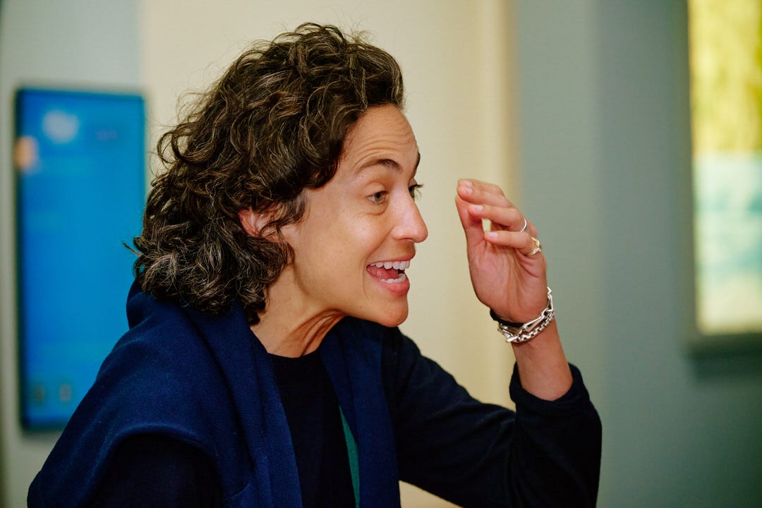 Woman with dark curly hair caught in profile mid-sentence in a hallway conversation, right hand raised with fingers together making her point, silver bracelets on her wrist