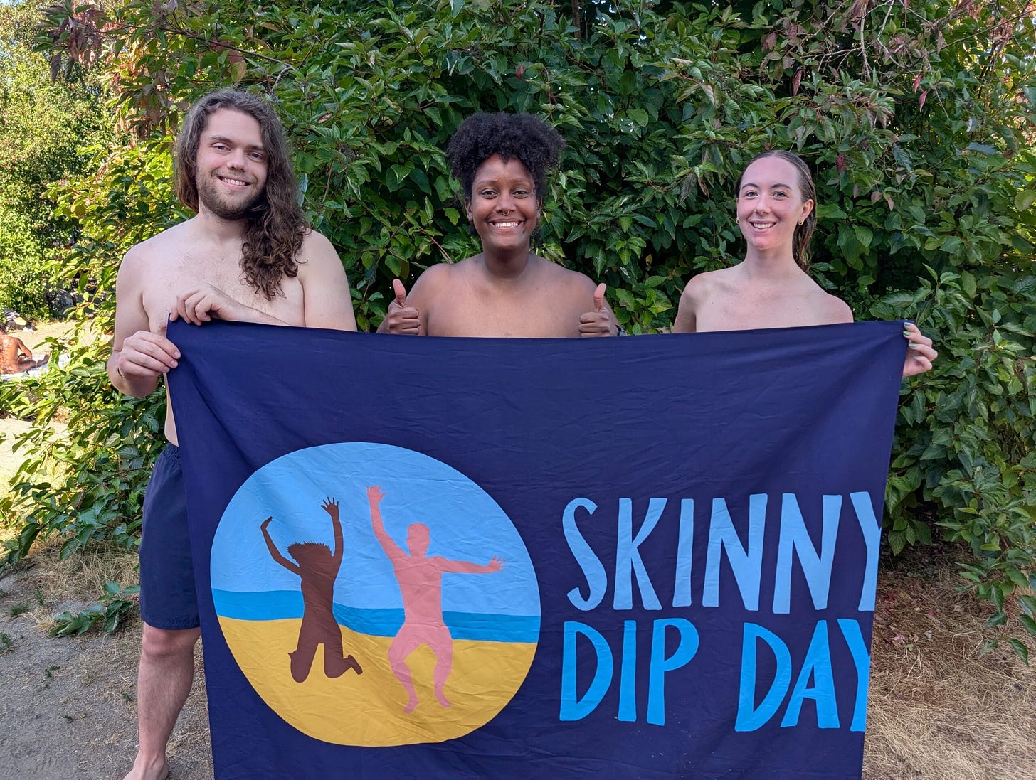 Three people from the Seattle Skinny Dippers group smile and pose nude behind a Skinny Dip Day flag, standing in front of lush greenery. Three people from the Seattle Skinny Dippers group smile and pose nude behind a Skinny Dip Day flag, standing in front of lush greenery.