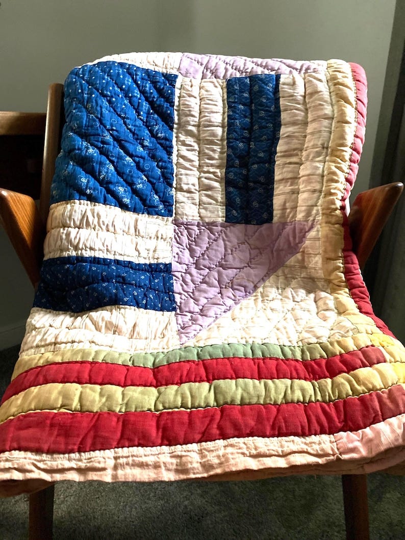 May include: A vintage quilt draped over a wooden chair. The quilt features a patchwork design with blue, white, red, and lavender sections. The blue sections have a floral pattern. The quilt is made of fabric and has a quilted texture. May include: A vintage quilt draped over a wooden chair. The quilt features a patchwork design with blue, white, red, and lavender sections. The blue sections have a floral pattern. The quilt is made of fabric and has a quilted texture.
