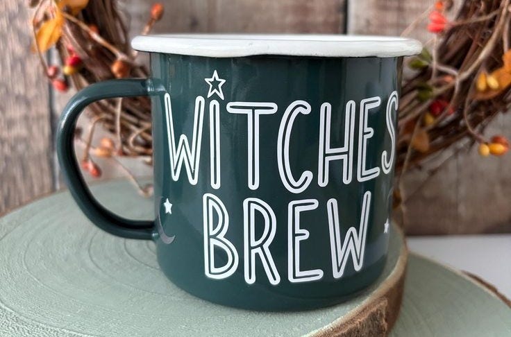 A dark green enamel mug with the words “WITCHES BREW” in bold white letters. The mug sits on a wood slice, surrounded by a rustic wreath with autumn leaves and berries — equal parts cosy and spellbinding.