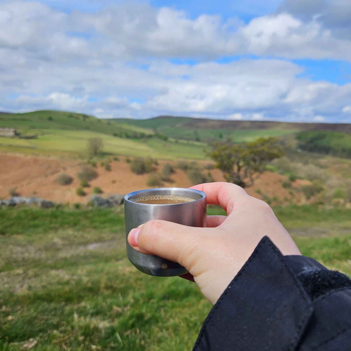 A hand holding a small metal cup filled with coffee. In the background are rolling green hills and a blue sky. A hand holding a small metal cup filled with coffee. In the background are rolling green hills and a blue sky.