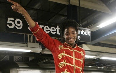 A young man faces camera with his right arm up and pointing to the side. He is wearing a red jacket and has curly hair.