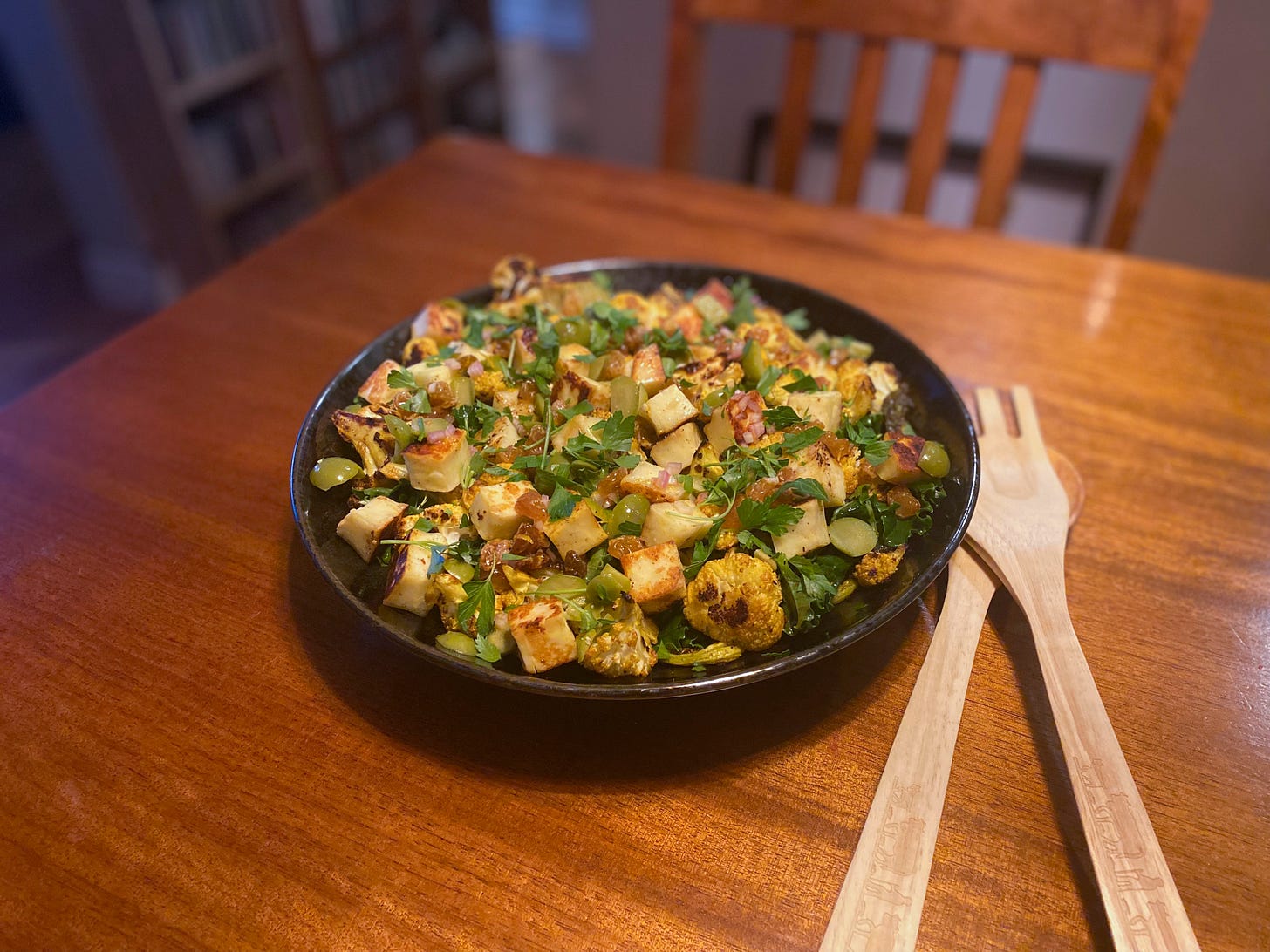 A shallow black serving dish filled with the salad described above: kale and parsley leaves, cubes of seared halloumi, pieces of green olive, and roasted browned florets of cauliflower. Wooden salad serving utensils sit on top of each other on the table beside it.