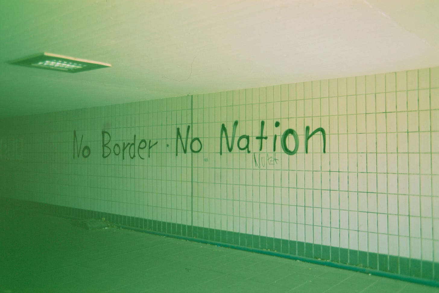 A green-tinged photo of what looks like a tile wall in a subway with "No Border - No Nation" graffitied onto it