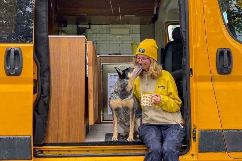 Scout the blue heeler and Haley the young woman sit on the stoop of a yellow converted campervan