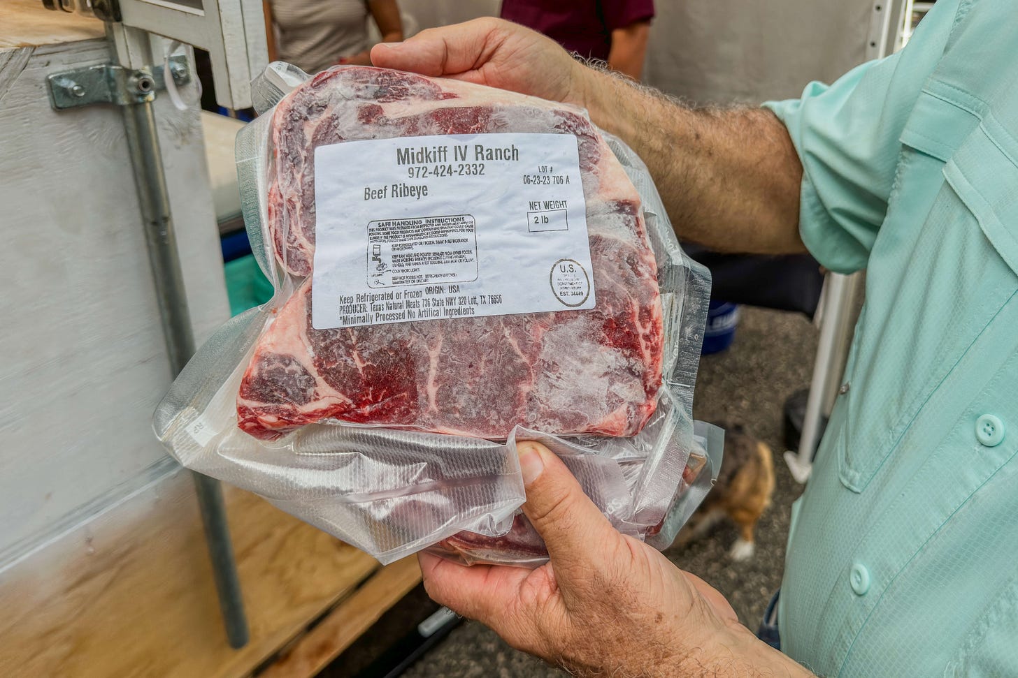 Ein Metzger hält auf dem New Braunfels Farmers Market ein vakuumverpacktes Rinder-Ribeye-Steak hoch. Auf dem Etikett steht Midkiff IV Ranch, USDA certified grass-fed Angus beef und das Gewicht von 2 Pfund, umgerechnet 900 Gramm, Die marmorierte Fleischstruktur ist durch die Verpackung sichtbar.