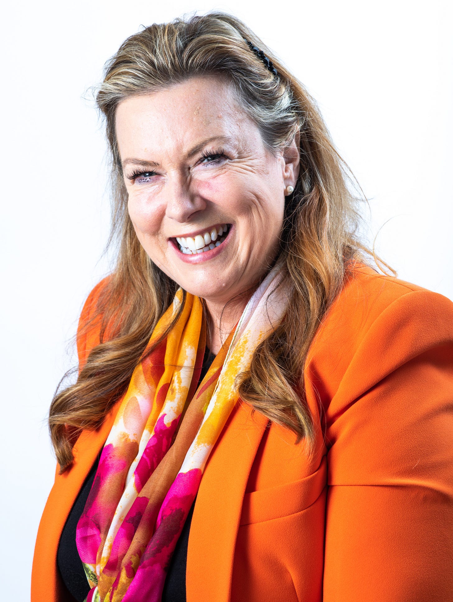 A headshot of Libby Hill. She is a white woman with long brown and grey hair, and she is smiling. She is wearing an orange jacket and an orange, pink and white scarf.