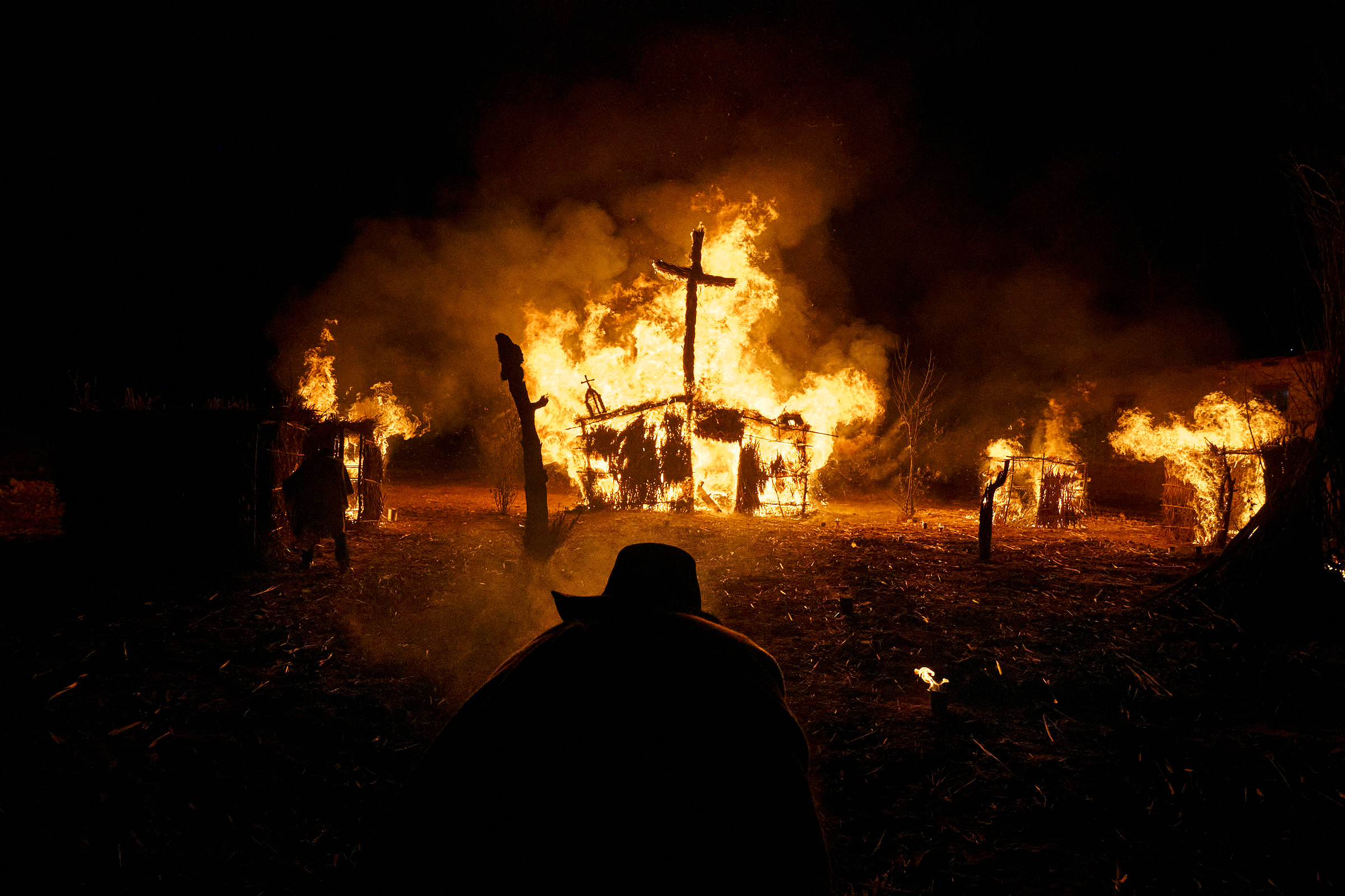 A person in a hat stands in the foreground, silhouetted against large flames consuming wooden structures and a cross at night. The scene is dramatic and intense, with fire lighting up the dark surroundings.