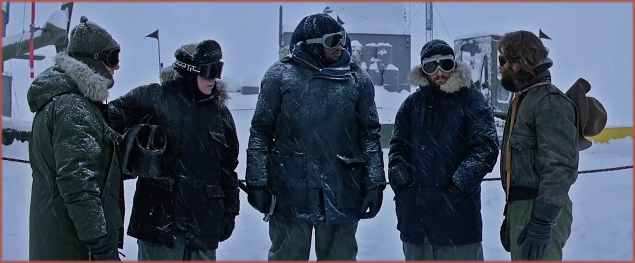 Film frame from The Thing (1982): a group of the researchers from Outpost 31 standing outside in snow, wearing dark parkas, goggles and gloves, a deliberately neutral wardrobe designed by Trish Keating to minimise individual identity and reinforce visual anonymity.