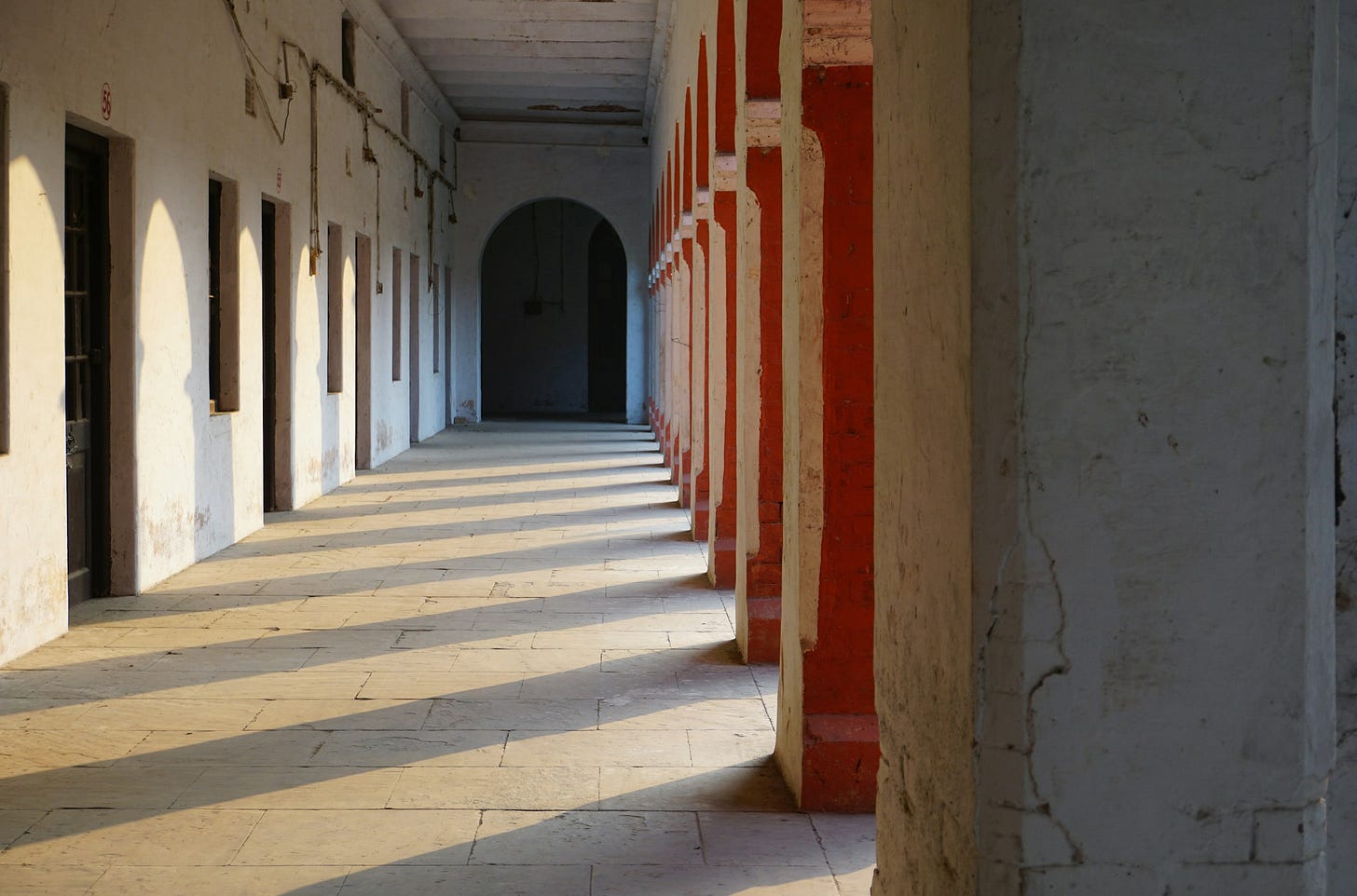 Un lungo portico coperto, probabilmente parte di un edificio storico o di un chiostro, inondato dalla luce del sole pomeridiano. Il corridoio è definito da una fila di archi rossi e pilastri bianchi a destra, che proiettano lunghe ombre oblique sul pavimento in pietra chiara. A sinistra, un muro bianco presenta una serie di aperture o porte scure che si susseguono in prospettiva. Sul fondo, il corridoio si restringe in un arco scuro, aumentando il senso di profondità.