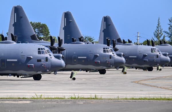 A line of large, gray military planes with propellers parked on an airfield tarmac. Their tails rise into a clear blue sky.