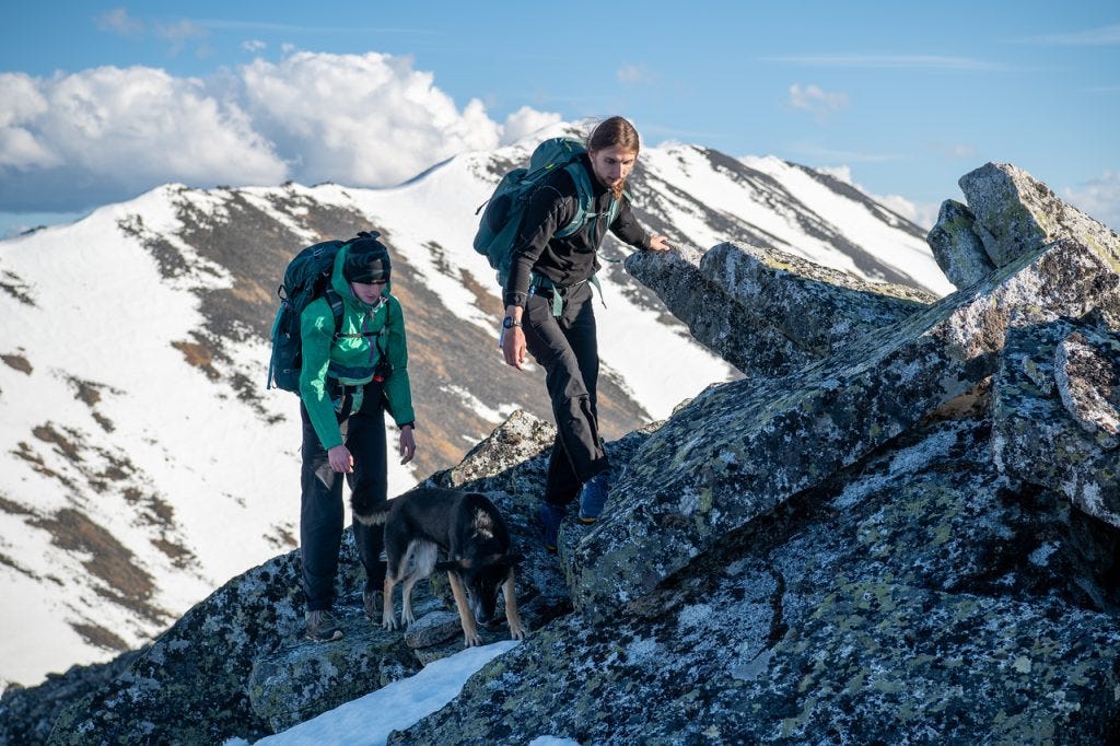 Two hikers and a black and tan dog pick their way across a jumble of lichen-covered granite blocks along a high ridge, snow-covered peaks behind.