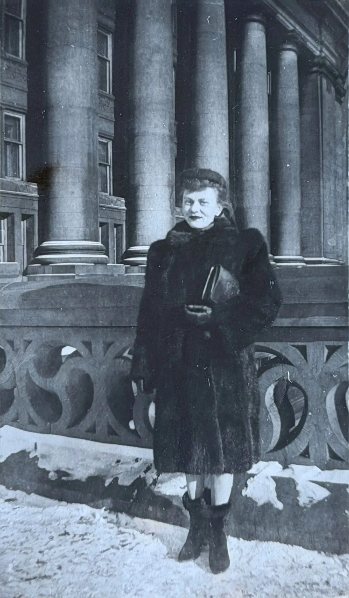 Photo ancienne en noir et blanc de Mamie Rose jeune, en long manteau de fourrure et chapeau, debout dans la neige devant un grand édifice à colonnes.