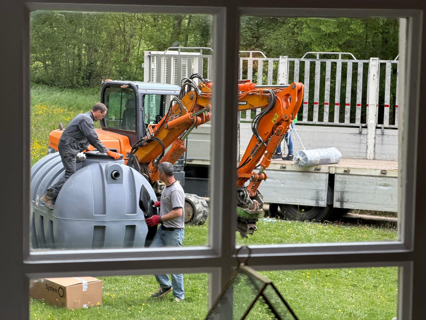 View through a wooden window frame of two workmen fitting a large grey plastic septic tank on a lawn, with an orange mini digger and a flatbed truck behind them, and a small glass terrarium hanging in the window in the foreground. View through a wooden window frame of two workmen fitting a large grey plastic septic tank on a lawn, with an orange mini digger and a flatbed truck behind them, and a small glass terrarium hanging in the window in the foreground.