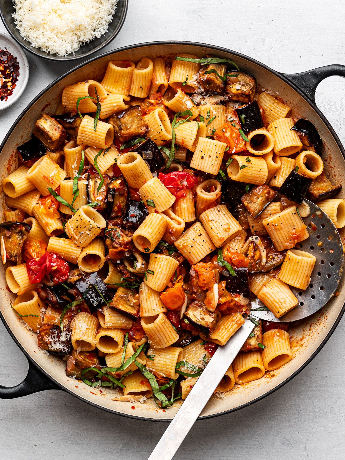 Roasted eggplant pasta in skillet with bowls of cheese and red pepper flakes on the side. Roasted eggplant pasta in skillet with bowls of cheese and red pepper flakes on the side.