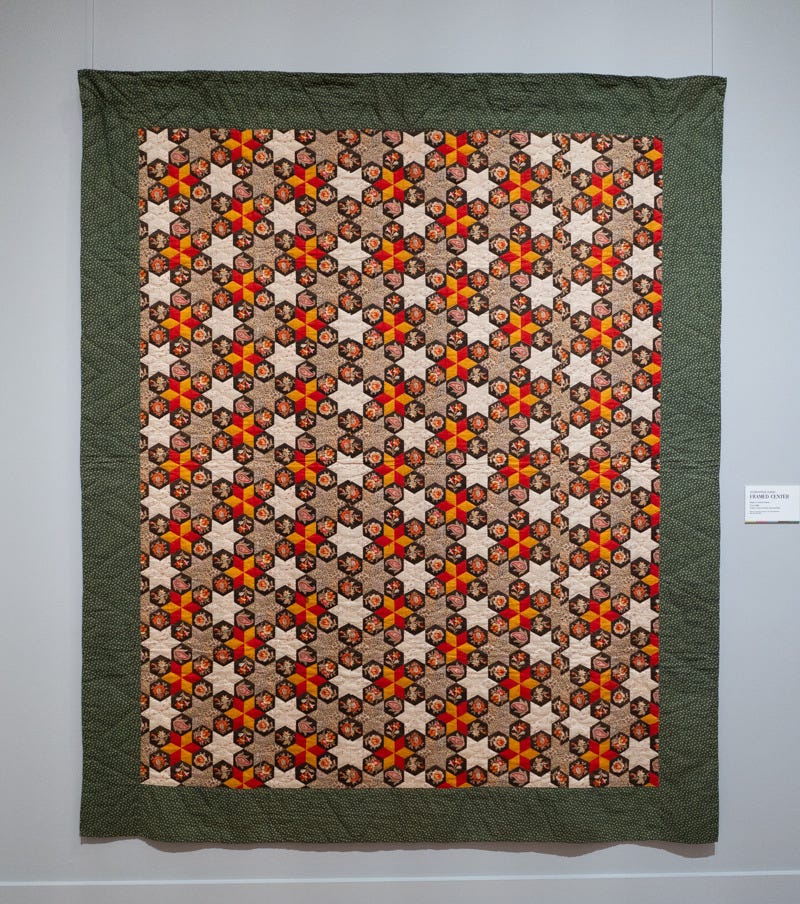 A photograph of an intricate patchwork quilt with a dark green border and an interior pattern of orange, yellow, and white stars A photograph of an intricate patchwork quilt with a dark green border and an interior pattern of orange, yellow, and white stars